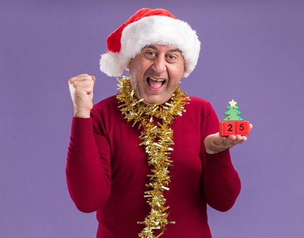 Hombre de mediana edad con gorro de Papá Noel de Navidad con oropel alrededor del cuello sosteniendo cubos de juguete con fecha veinticinco puño apretado feliz y emocionado de pie sobre fondo púrpura