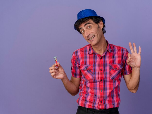 Hombre de mediana edad impresionado con gorro de fiesta sosteniendo un soplador de fiesta mirando al frente haciendo el signo de ok aislado en la pared púrpura