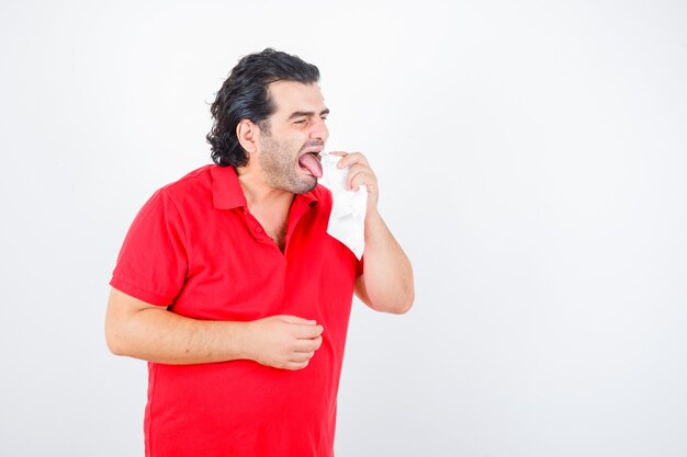 Hombre de mediana edad limpiando la lengua con una servilleta en camiseta roja y mirando disgustado, vista frontal.