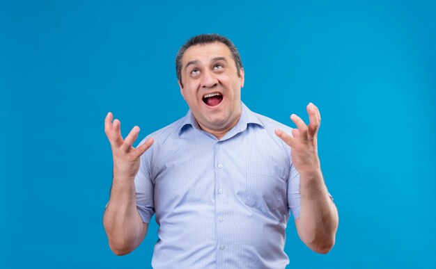 Hombre de mediana edad llorando y enojado con camisa de rayas azules gritando las manos en un espacio azul