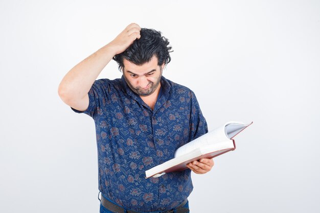 Hombre de mediana edad mirando a través del libro mientras se rasca la cabeza en camisa y mira pensativo, vista frontal.