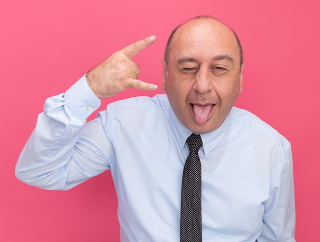 Hombre de mediana edad parpadeó alegre con camiseta blanca con corbata mostrando gesto de lengua y cabra aislado en la pared rosa