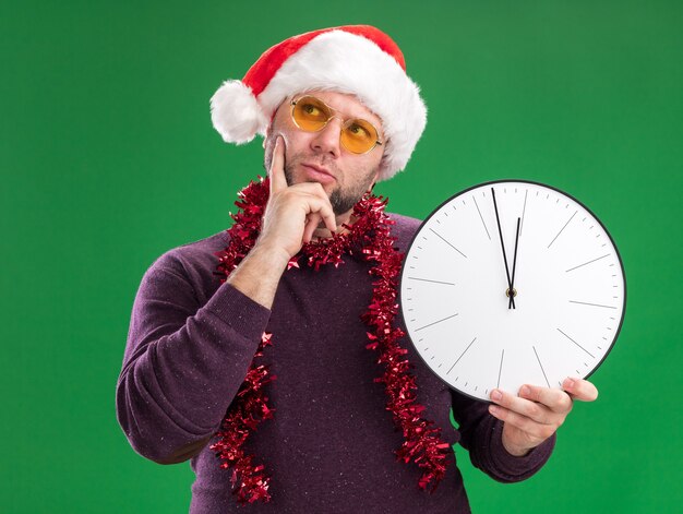 Hombre de mediana edad pensativo con gorro de Papá Noel y guirnalda de oropel alrededor del cuello con gafas sosteniendo el reloj manteniendo la mano en la barbilla mirando hacia arriba aislado en la pared verde