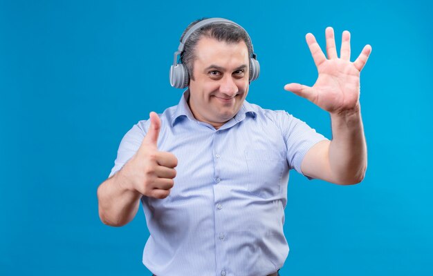 Hombre de mediana edad positivo y alegre en camisa de rayas azules usando audífonos mostrando con los dedos el número seis sobre un fondo azul.