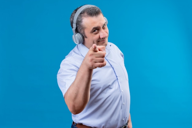 Hombre de mediana edad positivo en camisa de rayas azules usando audífonos apuntando con el dedo índice a la cámara sobre un fondo azul.
