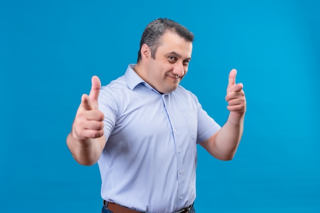 Hombre de mediana edad positivo y sonriente en camisa de rayas azules apuntando con el dedo índice a la cámara sobre un fondo azul.