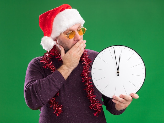 Hombre de mediana edad preocupado con gorro de Papá Noel y guirnalda de oropel alrededor del cuello con gafas manteniendo la mano en la boca sosteniendo y mirando el reloj aislado en la pared verde