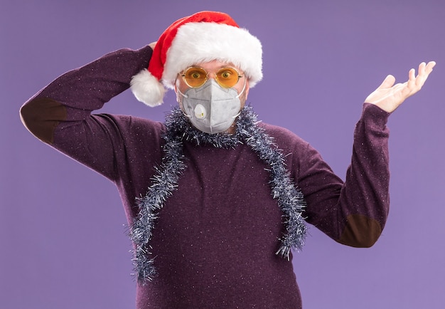 Hombre de mediana edad preocupado con gorro de Papá Noel y guirnalda de oropel alrededor del cuello con gafas y máscara protectora mirando a la cámara mostrando la mano vacía manteniendo la mano detrás de la cabeza aislada sobre fondo púrpura