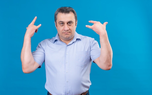 Hombre de mediana edad con problemas en la camisa a rayas azules tomados de la mano en el símbolo de la roca sobre un fondo azul.