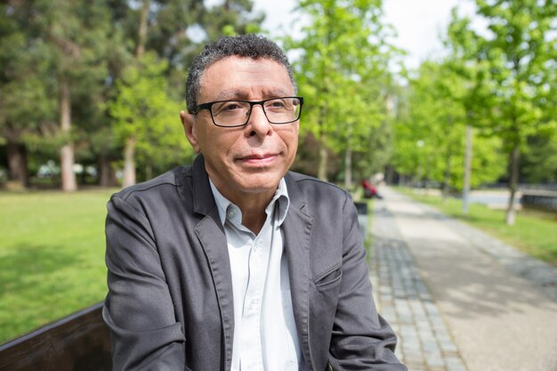 Foto gratuita hombre de mediana edad relajado que se sienta en banco en parque de la ciudad