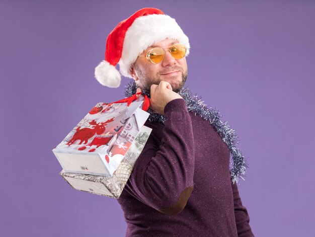 Hombre de mediana edad satisfecho con gorro de Papá Noel y guirnalda de oropel alrededor del cuello con gafas de pie en vista de perfil sosteniendo bolsas de regalo de Navidad en el hombro mirando a cámara aislada sobre fondo púrpura