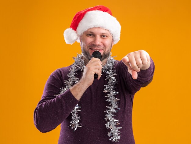 Hombre de mediana edad satisfecho con gorro de Papá Noel y guirnalda de oropel alrededor del cuello hablando por el micrófono mirando y apuntando a la cámara aislada sobre fondo naranja