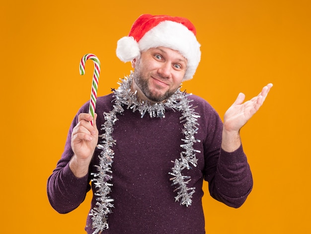 Hombre de mediana edad satisfecho con gorro de Papá Noel y guirnalda de oropel alrededor del cuello sosteniendo bastón dulce de Navidad mirando a cámara mostrando la mano vacía aislada sobre fondo naranja