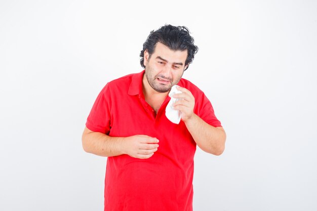 Hombre de mediana edad secándose los ojos con una servilleta mientras llora en camiseta roja y mirando ofendido, vista frontal.