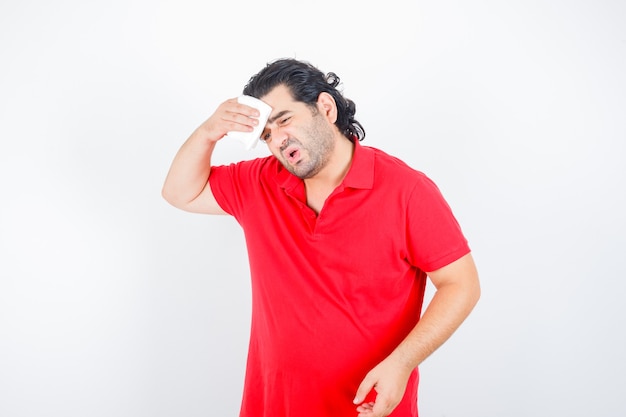 Hombre de mediana edad secándose el sudor en camiseta roja y mirando enfermo, vista frontal.