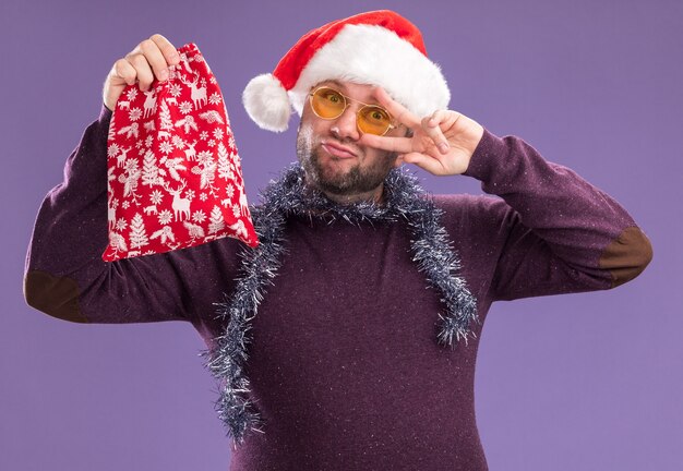 Hombre de mediana edad seguro con gorro de Papá Noel y guirnalda de oropel alrededor del cuello con gafas sosteniendo un saco de regalo de Navidad mirando a la cámara mostrando el símbolo de signo v cerca del ojo aislado sobre fondo púrpura