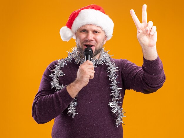 Hombre de mediana edad seguro con gorro de Papá Noel y guirnalda de oropel alrededor del cuello hablando por el micrófono haciendo el signo de la paz aislado en la pared naranja