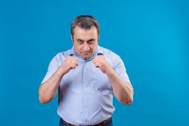 Hombre de mediana edad serio y enojado vestido con camisa de rayas verticales azul practicando movimientos de boxeo sobre un fondo azul.