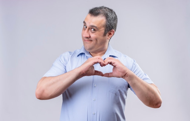 Hombre de mediana edad sonriente en camisa azul a rayas verticales que muestra el signo del corazón con las manos mientras está de pie sobre un fondo blanco.