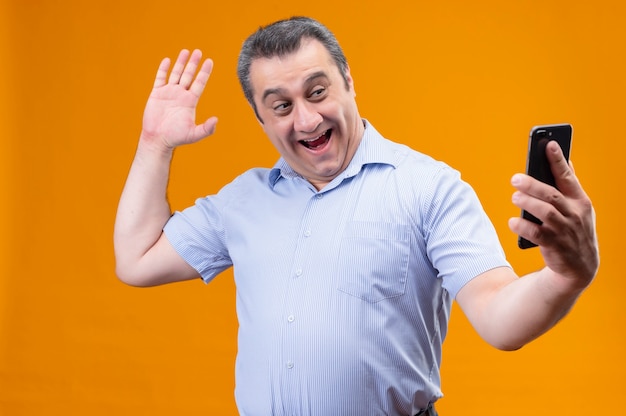Hombre de mediana edad sonriente y positivo en auriculares de camisa a rayas azules agitando la mano y saludando mientras habla por videollamada usando su teléfono inteligente mientras está de pie sobre un fondo naranja