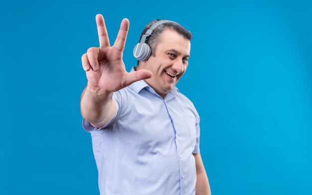 Hombre de mediana edad sonriente y positivo en camisa de rayas azules en auriculares mostrando con los dedos el número seis sobre un fondo azul.