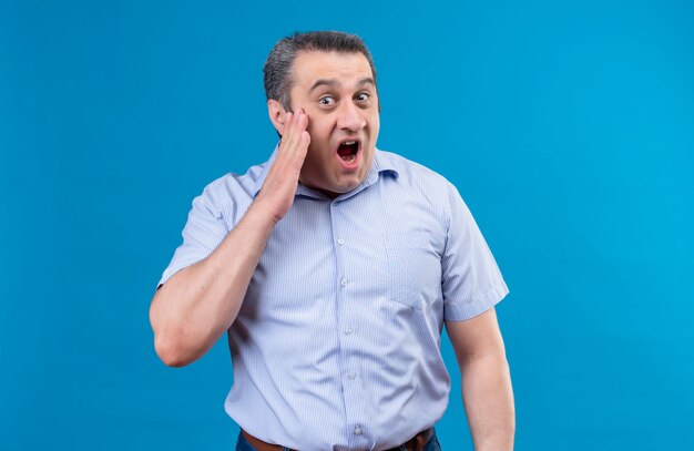 Hombre de mediana edad sorprendente y asombrado en camisa azul manteniendo la boca abierta sosteniendo la mano en la mejilla sobre un fondo azul