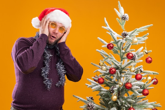 Hombre de mediana edad sorprendido con gorro de Papá Noel y guirnalda de oropel alrededor del cuello con gafas de pie cerca del árbol de Navidad decorado manteniendo las manos en la cabeza aislada en la pared naranja