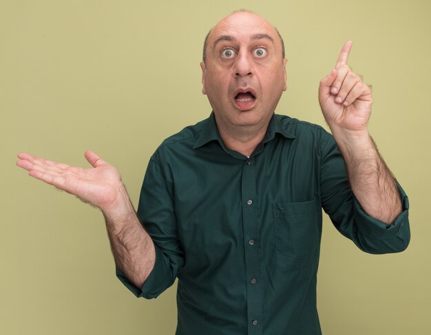 Hombre de mediana edad sorprendido con puntos de camiseta verde con la mano en los puntos laterales en arriba aislado en la pared verde oliva