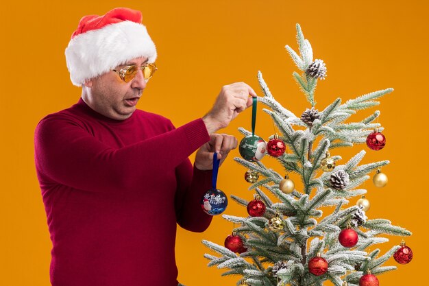 Hombre de mediana edad sorprendido vestido con gorro de Papá Noel de Navidad en cuello alto rojo oscuro y gafas amarillas que decoran el árbol de Navidad de pie sobre la pared naranja