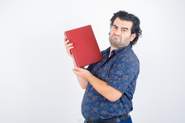 Hombre de mediana edad sosteniendo el libro sobre el pecho en camisa y mirando vacilante, vista frontal.