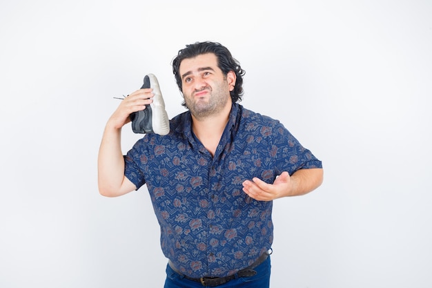 Hombre de mediana edad sosteniendo el zapato sobre el hombro mientras estira la mano en gesto de interrogación en camisa y mira vacilante, vista frontal.