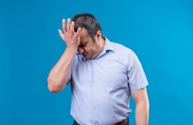 Hombre de mediana edad triste y deprimido en camisa azul a rayas verticales sosteniendo la mano en la frente en un espacio azul