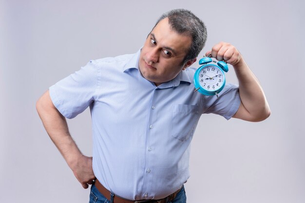 Hombre de mediana edad vestido con camisa azul de rayas verticales escuchando el tictac del reloj sosteniendo el reloj despertador azul sobre un fondo blanco.