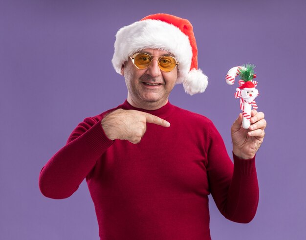 Hombre de mediana edad vestido con gorro de Papá Noel de Navidad en gafas amarillas sosteniendo bastón de caramelo de Navidad apuntando con el dedo índice sonriendo de pie sobre fondo púrpura