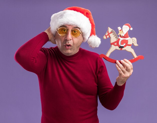 Hombre de mediana edad vestido con gorro de Papá Noel de Navidad en gafas amarillas sosteniendo juguetes de Navidad mirando a cámara asombrado y sorprendido de pie sobre fondo púrpura