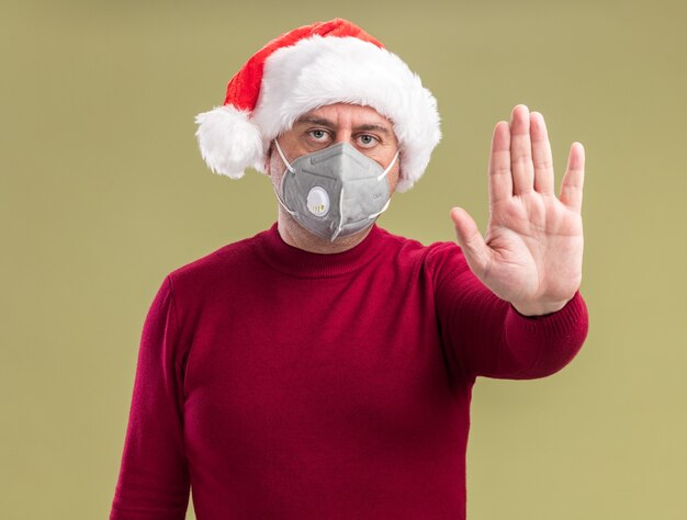 Hombre de mediana edad vestido con gorro de Papá Noel de Navidad con máscara protectora facial haciendo gesto de parada con la mano con cara seria de pie sobre la pared verde