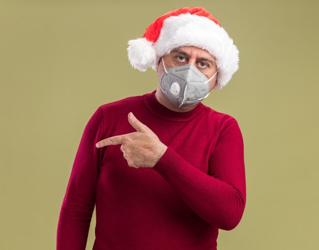 Hombre de mediana edad vestido con gorro de Papá Noel de Navidad con máscara protectora facial mirando a la cámara con cara seria apuntando con el dedo índice hacia el lado de pie sobre fondo verde