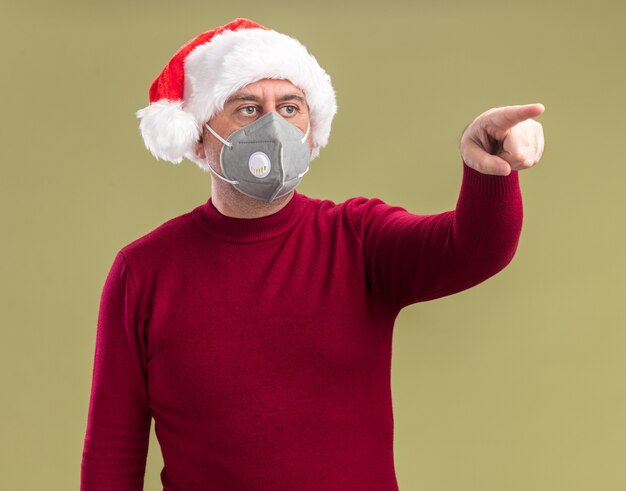 Hombre de mediana edad vestido con gorro de Papá Noel de Navidad con máscara protectora facial mirando a un lado con expresión de preocupación apuntando con el dedo índice hacia el lado de pie sobre fondo verde