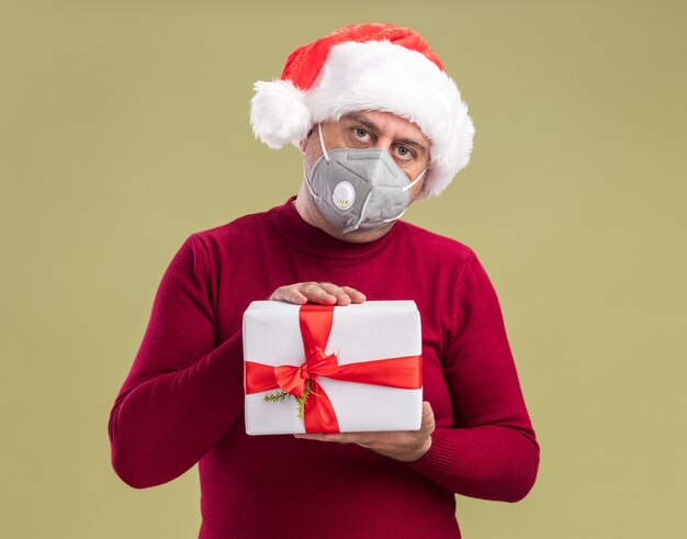 Hombre de mediana edad vestido con gorro de Papá Noel de Navidad con máscara protectora facial sosteniendo el regalo de Navidad con cara seria de pie sobre la pared verde