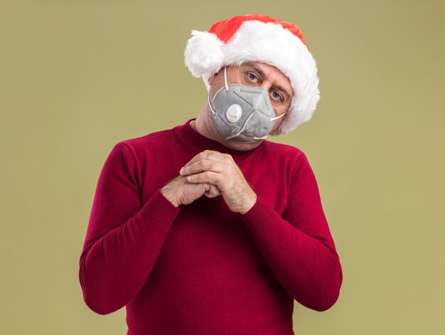 Hombre de mediana edad vestido con gorro de Papá Noel de Navidad con máscara protectora facial tomados de la mano junto con la cara seria de pie sobre la pared verde