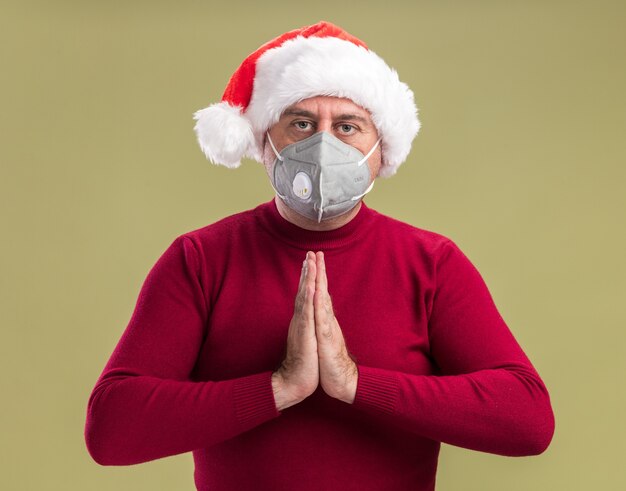 Hombre de mediana edad vestido con gorro de Papá Noel de Navidad con máscara protectora facial tomados de la mano juntos como rezando con cara seria de pie sobre fondo verde