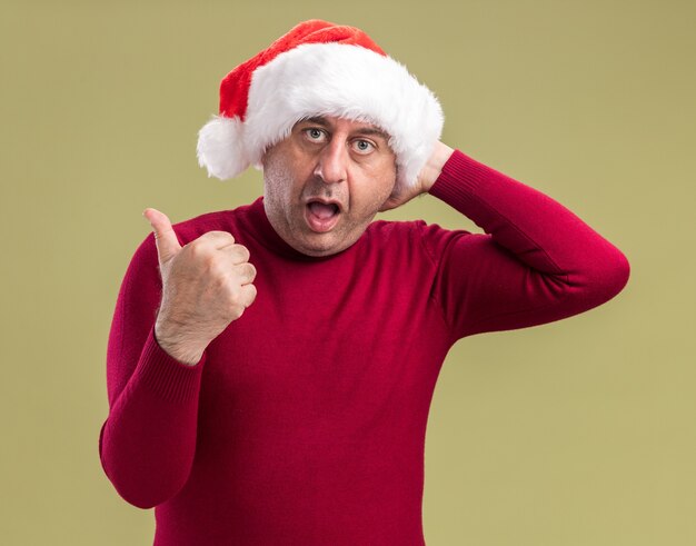 Hombre de mediana edad vestido con gorro de Papá Noel de Navidad mirando a la cámara confundido apuntando con el pulgar hacia el lado de pie sobre fondo verde