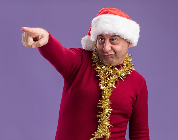 Hombre de mediana edad vestido con gorro de Papá Noel de Navidad con oropel alrededor del cuello apuntando con el índice figner hacia el lado confundido y muy ansioso de pie sobre fondo púrpura