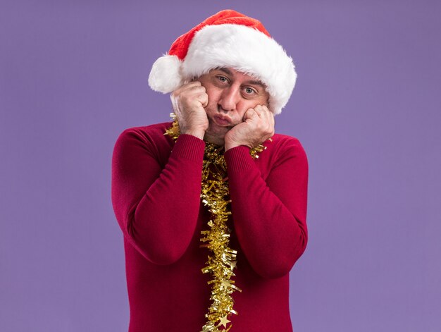 Hombre de mediana edad vestido con gorro de Papá Noel de Navidad con oropel alrededor del cuello mirando a la cámara con confundir expresión de mejillas de pie sobre fondo púrpura