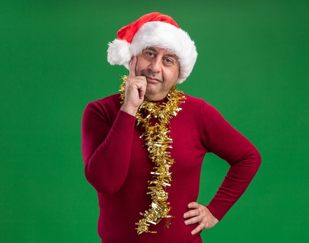 Hombre de mediana edad vestido con gorro de Papá Noel de Navidad con oropel alrededor del cuello mirando a la cámara sonriendo confiados de pie sobre fondo verde