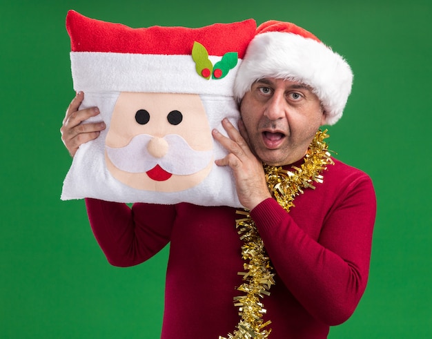 Foto gratuita hombre de mediana edad vestido con gorro de papá noel de navidad con oropel alrededor del cuello sosteniendo almohada de navidad mirando a la cámara asombrado y sorprendido de pie sobre fondo verde