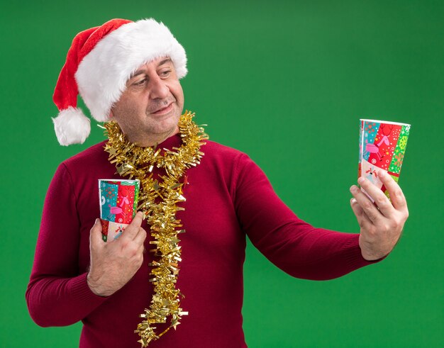 Hombre de mediana edad vestido con gorro de Papá Noel de Navidad con oropel alrededor del cuello sosteniendo coloridos vasos de papel mirando confundido tener dudas sobre fondo verde