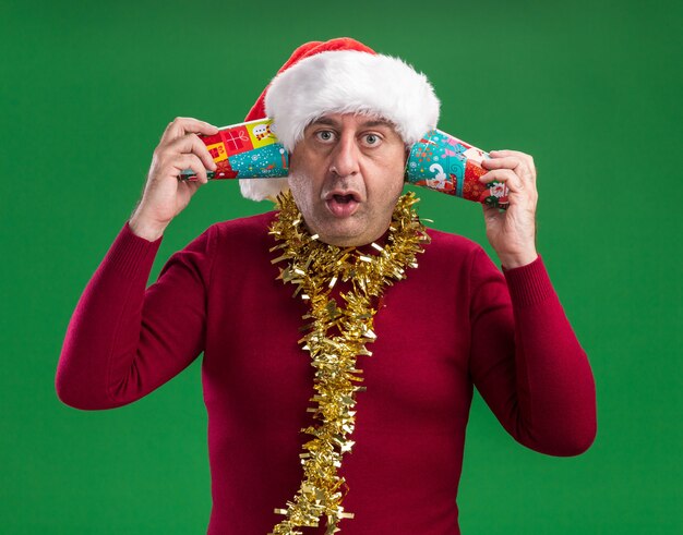 Hombre de mediana edad vestido con gorro de Papá Noel de Navidad con oropel alrededor del cuello sosteniendo coloridos vasos de papel