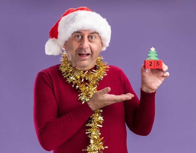 Hombre de mediana edad vestido con gorro de Papá Noel de Navidad con oropel alrededor del cuello sosteniendo cubos de juguete con fecha veinticinco presentando con el brazo de la mano sonriendo de pie sobre fondo púrpura