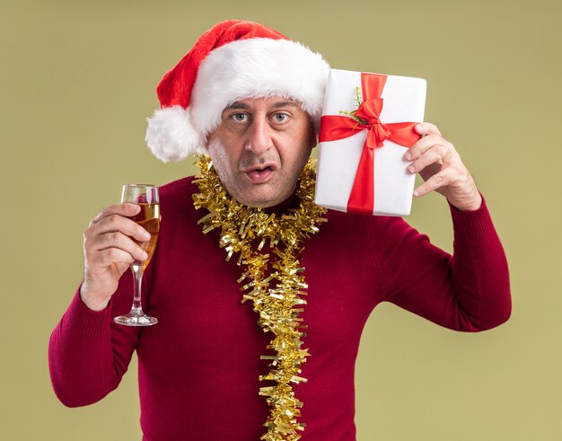 Hombre de mediana edad vestido con gorro de Papá Noel de Navidad con oropel alrededor del cuello sosteniendo el regalo de Navidad y una copa de champán mirando a la cámara confundido de pie sobre fondo verde